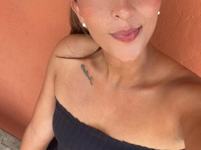 EmelyOwen's upper body, black strapless top, small tits, tattoo on right shoulder, pink lips, slightly parted, pearl earrings, light brown hair, orange wall background, warm skin tone.