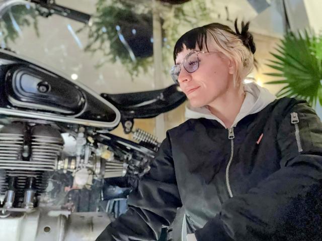 SaraleePoison, con gafas, una chaqueta negra y el cabello rubio recogido en un moño, apoyada en una motocicleta negra con un motor brillante. Está mirando hacia un lado, sonriendo ligeramente..