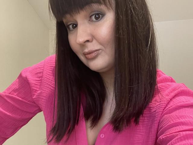 AlinaMilf has straight brown hair, bangs, and a pink ribbed shirt. She's smiling with brown eyes and light skin. Her small tits are partially visible.