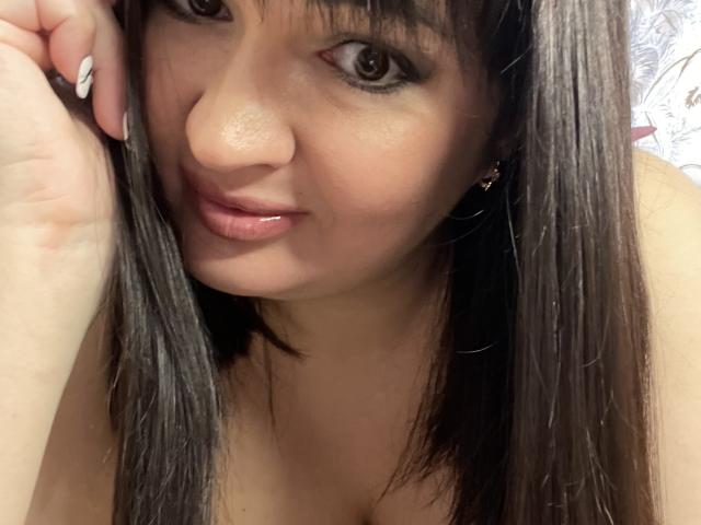 AlinaMilf has straight brown hair, brown eyes, and a cute smile with glossy pink lips. She's touching her hair with her hand, showing off her manicured nails.