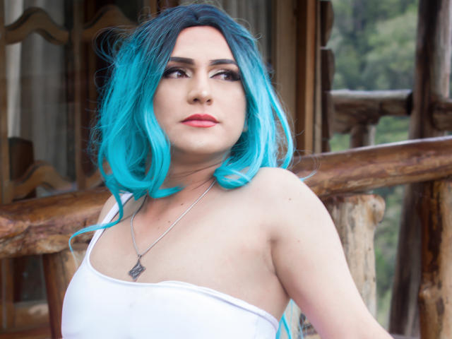 Iris Durand, con i capelli blu brillante, un top bianco senza spalline e una collana d'argento, guarda di lato con un'espressione seria, mostrando le spalle scoperte e un seno piccolo..