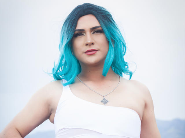 IrisDurand with bright blue, wavy hair, white one-shoulder top, and silver necklace, showing a confident, sexy, cute face with pink lipstick, and a subtle smile.