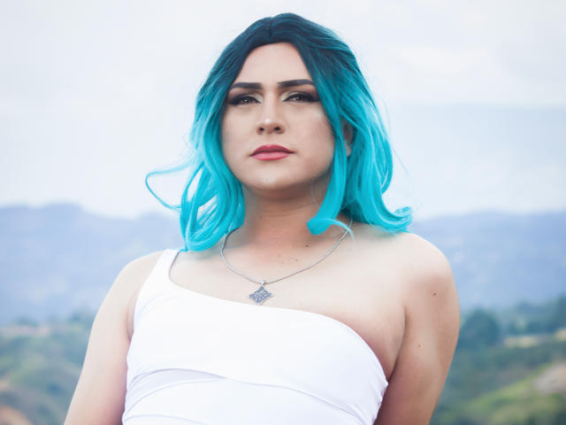 IrisDurand with bright blue hair, white one-shoulder top, and silver necklace, looking serious. Boobs partially visible. Pale skin, dark eyebrows, pink lips. Blue-gray mountains blurred in background.