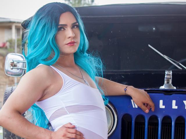IrisDurand with bright blue hair, wearing a white tank top, leaning on a blue Jeep, showing a slim, flat-ass, and a small, cute, unobstructed pussy.