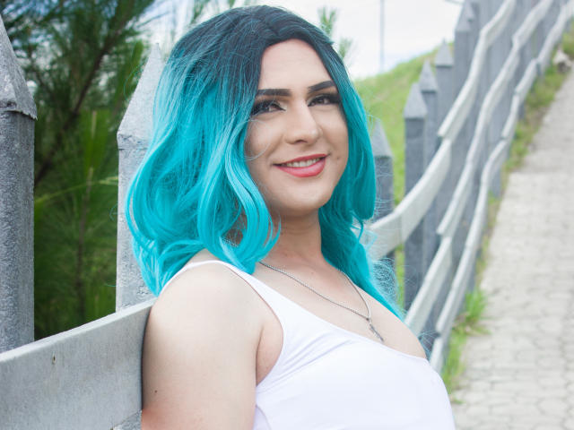 Iris Durand, avec ses cheveux bleu vif, souriante, portant un débardeur blanc, appuyée contre une clôture grise, montrant ses seins, debout sur un chemin de gravier, avec du vert en arrière-plan..