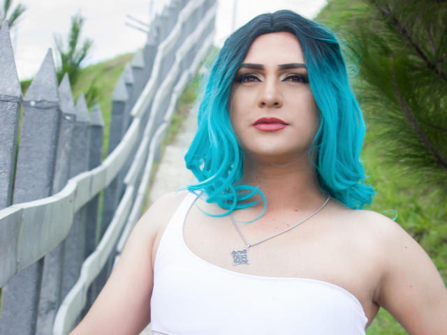 IrisDurand with bright blue hair, white tank top, and silver necklace, standing by a gray fence, showing off her tits and a serious face, green foliage in background.