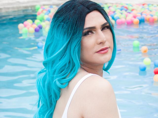 IrisDurand, avec ses cheveux bleu turquoise, son débardeur blanc et son rouge à lèvres rose, regarde par-dessus son épaule vers l'appareil photo, dans une piscine remplie de balles colorées. Les seins d'IrisDurand sont visibles..