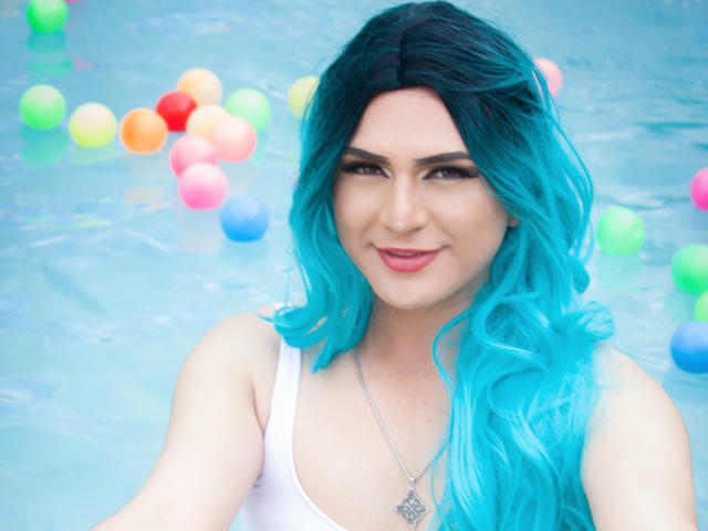 Iris Durand, avec ses cheveux bleu vif, son débardeur blanc, souriante, avec une poitrine de taille moyenne, dans une piscine remplie de balles colorées, portant un collier en argent avec un pendentif en forme de diamant..