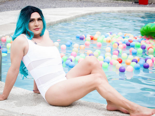IrisDurand with blue hair, white one-piece, sitting by a pool filled with colorful balls, legs crossed, arms behind, looking at camera, smiling, legs up, pool edge.