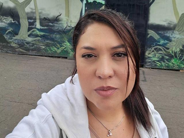 PaulaAnd has a pouty face, brown eyes, and medium brown skin. She's wearing a white hoodie, has dark brown hair in a ponytail, and a silver necklace with a pendant.