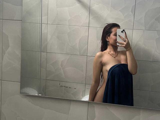 VannesaSaenz with wet long dark hair, naked except for a dark blue towel covering her ass, taking a mirror selfie with her phone, showing her tits and smiling.