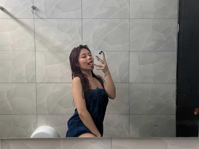 VannesaSaenz with wet dark hair, tongue out, taking a mirror selfie with a phone, wearing a dark blue towel, showing her ass, in a tiled bathroom.