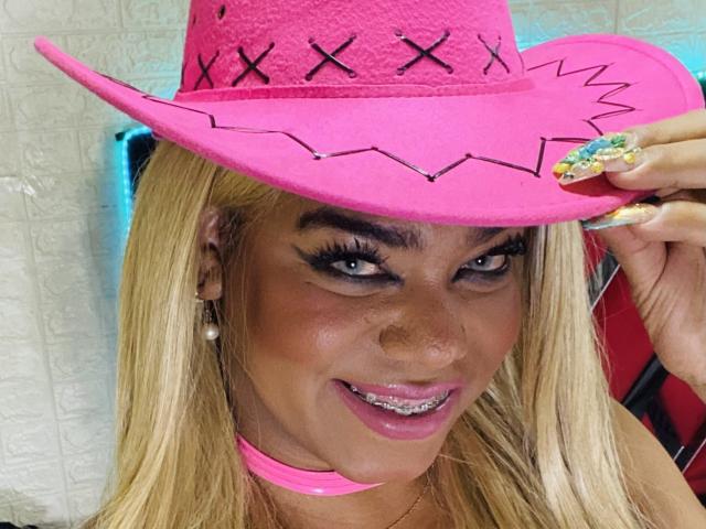 NatashaBurgss indossa un cappello da cowboy rosa acceso con delle "X" nere, tiene il cappello con le unghie scintillanti, sorride mostrando i denti, ha i capelli biondi, una collana rosa e sopracciglia scure..