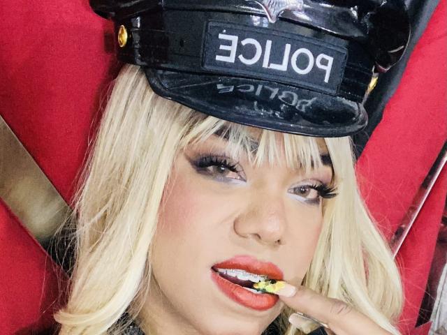 NatashaBurgss with blonde hair, wearing a black police cap, has red lipstick, yellow nails, and is sucking a yellow lollipop, with heavy makeup and a seductive expression.