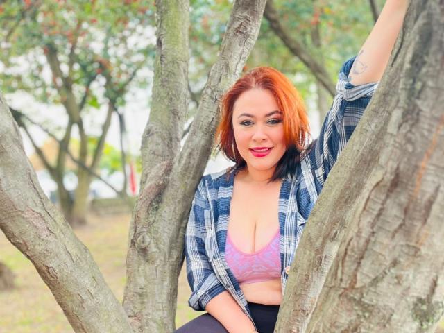 MaddyBlooms with red hair, wearing an unbuttoned plaid shirt, exposing her pink lace bra and showing her tits, arm raised, standing between tree trunks, smiling.