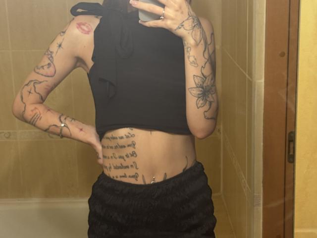SensualMila's tattooed arms, black crop top, black pants, belly button piercing, and hand on hip in a bathroom mirror selfie. Her tits are covered, but her ass is not.