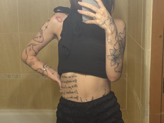 SensualMila with tattoos, black sleeveless top, and black pants, showing her slim, flat-assed, and pierced belly in a bathroom mirror selfie. Her arms have flowers and script.