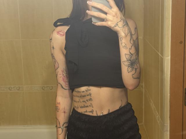 SensualMila, avec des tatouages, portant un haut noir sans manches et un pantalon noir, tenant un téléphone dans un selfie devant un miroir, montrant son ventre plat et ses tatouages..