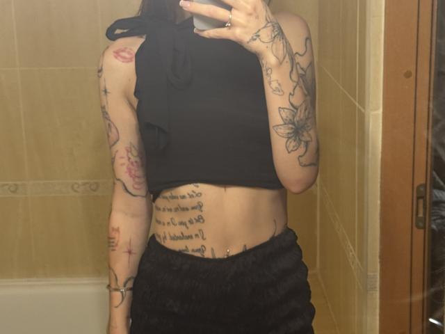 SensualMila con tatuajes en los brazos y las costillas, top corto negro, pantalones negros, selfie en el espejo, sosteniendo el teléfono, cabello negro largo, delgada, con glúteos visibles, tatuaje de flor en el brazo derecho..