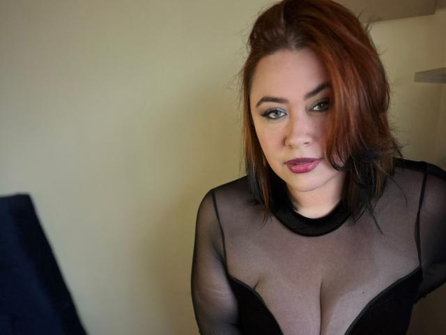 MaddyBlooms with red-black hair, wearing sheer black top, showing her tits, has green eyes, pink lipstick, and a serious look, sitting in front of a computer.