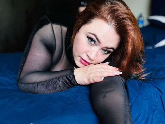 MaddyBlooms with red hair, wearing black sheer top and tights, lying on blue bed, head on arm, looking at camera, slight smile, blue eyes, red lipstick, sexy.