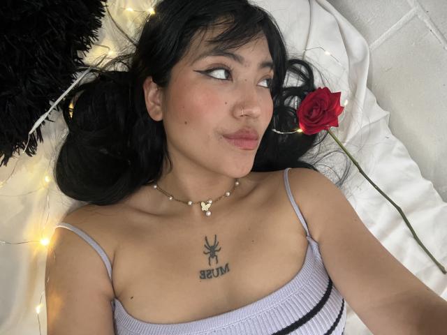 Elizaa with black hair, white eyeliner, pouty lips, small tits, tattooed spider on chest, wearing purple top, necklace with butterfly, lying on bed with red rose and string lights.