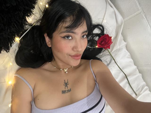 Elizaa with black hair, wearing a white tank top, lying on a bed, smiling, red rose by her cheek, "SEED" tattoo on chest, fairy lights, black pillow.
