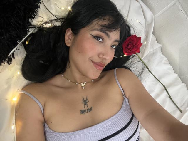 Elizaa with black hair, wearing a white ribbed top, lying on a bed with string lights, red rose, and black pillow, showing a spider tattoo above her tits.