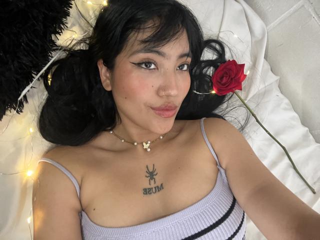 Elizaa with black pigtails, a red rose, and fairy lights, lying on a white bed, wearing a spaghetti strap top, showing her small tits and a "SEIN" tattoo.