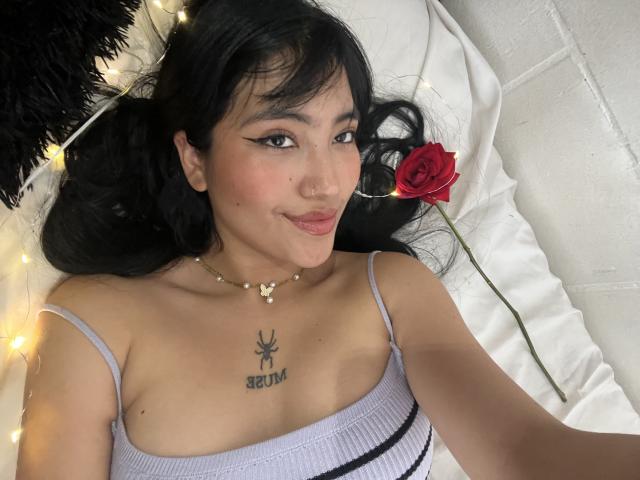 Elizaa with black hair, wearing a white tank top, lying down, rose in hand, string lights, butterfly necklace, bee tattoo under tits, subtle smile, nose ring.