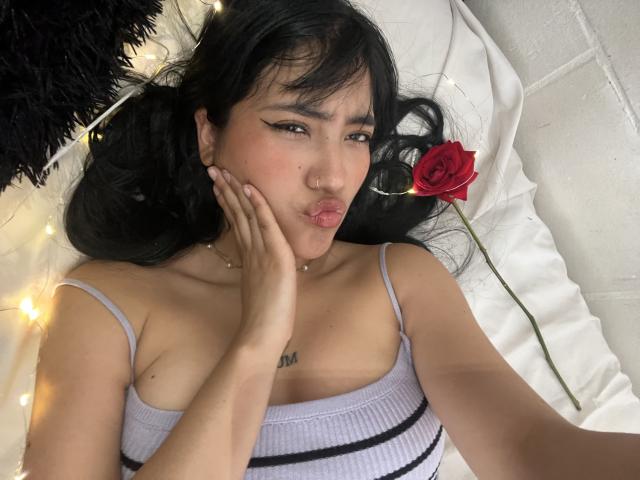 Elizaa with black hair, pouty lips, and a nose ring, wearing a striped top, touching her cheek, and a red rose beside her. Fairy lights in the background.