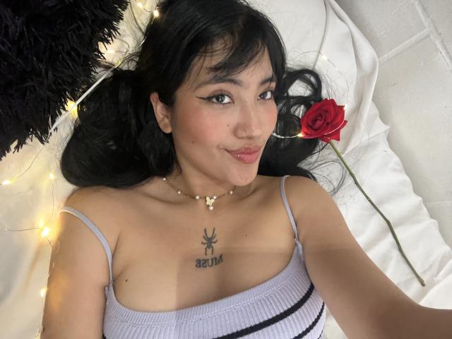 Elizaa with black hair, wearing a white tank top, lying down, smiling, rose next to her, small gold necklace, weed tattoo above her tits, string lights in background.