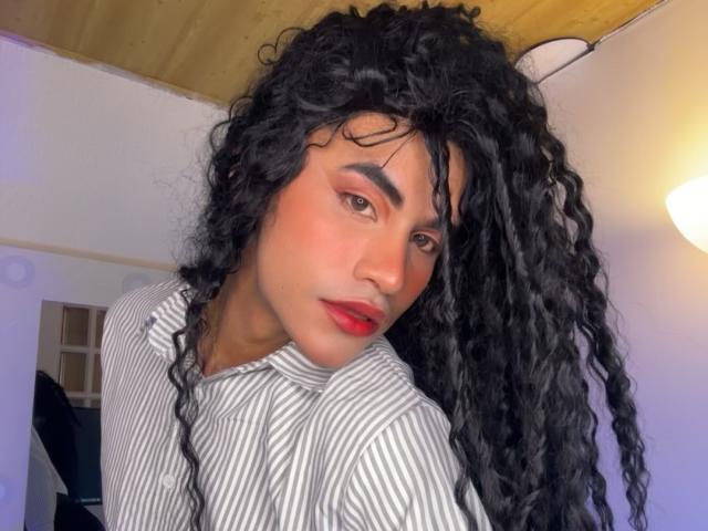 HannielVega with curly black hair, red lipstick, and striped shirt, looking at camera, brown eyes, slight smile, wood ceiling, white walls, ceiling light.