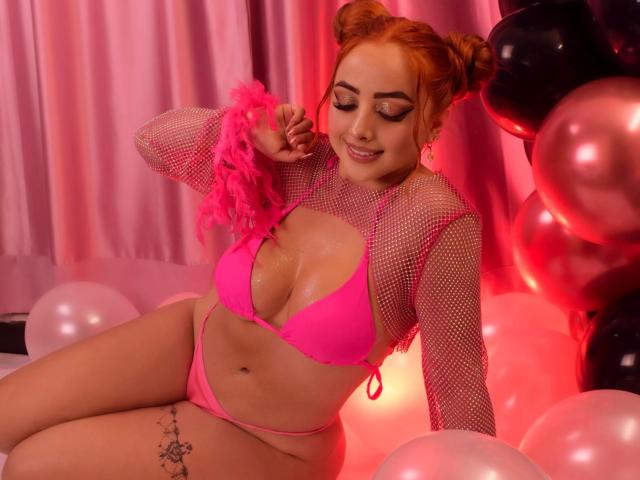KylieCharm with red hair in pink bikini, fishnet top, holding pink feather, sitting on balloons, smiling, showing her shiny tits, tattoo on right thigh, pink and black balloons.