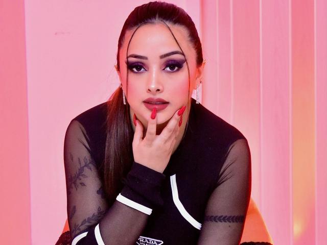 KylieCharm with red lipstick, black sheer top, tattoos on arms, red nails, hand on chin, pink and orange lighting, long brown ponytail, and serious expression.