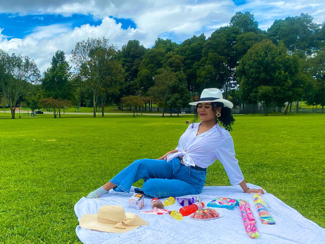SophieSonyas sitting on a white blanket with a green lawn, wearing a white shirt, blue jeans, and a white hat, with colorful snacks and a straw hat in front.