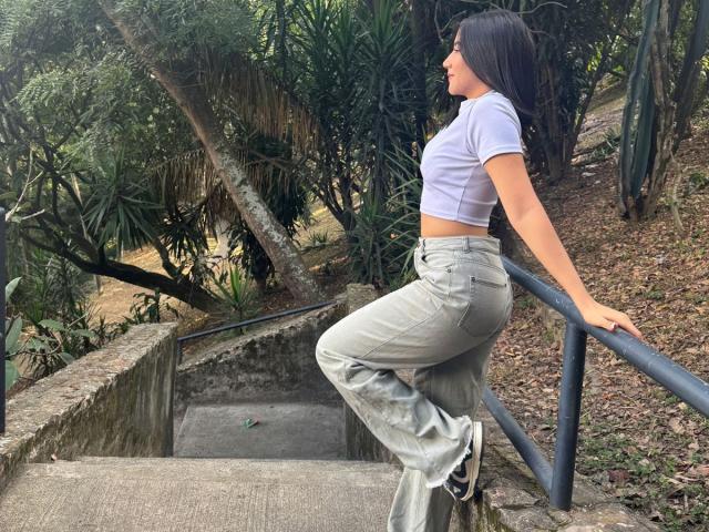 JuanaFox with long black hair, white crop top, and gray pants, leaning on a metal railing, showing her ass, standing on concrete stairs in a wooded area.