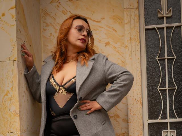 RoxieJackson with red hair, glasses, grey coat, black lingerie with gold trim, showing her tits, hand on wall, standing near a metal door.