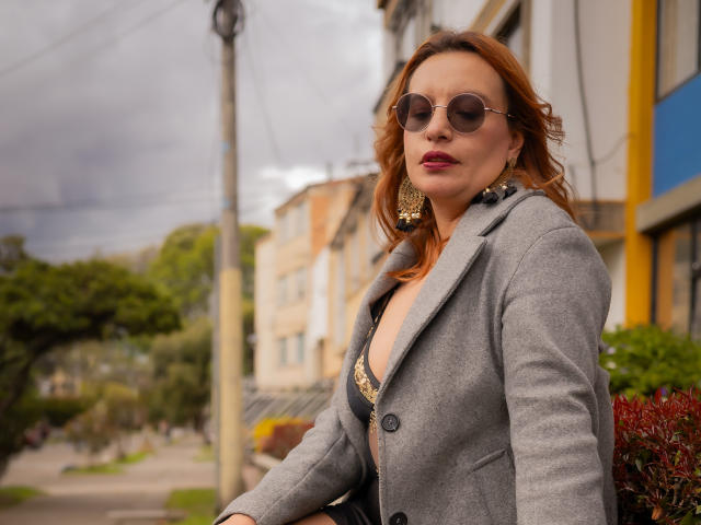 RoxieJackson with wavy red hair, wearing round sunglasses, grey coat, black bra with gold trim, and large earrings, standing outside with a confident, slightly smoky look.