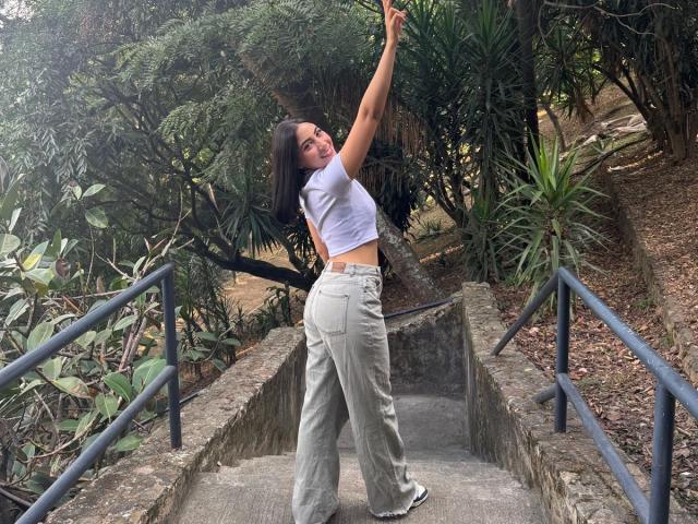 JuanaFox with medium tits, wearing a white crop top and high-waisted jeans, stands on concrete stairs, arms raised, smiling, with green foliage and metal railings in the background.