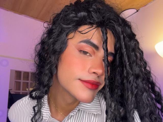 HannielVega has curly black hair, red lipstick, and a gold necklace, wearing a striped shirt with pink eyeshadow and a slight smile, looking at the camera.