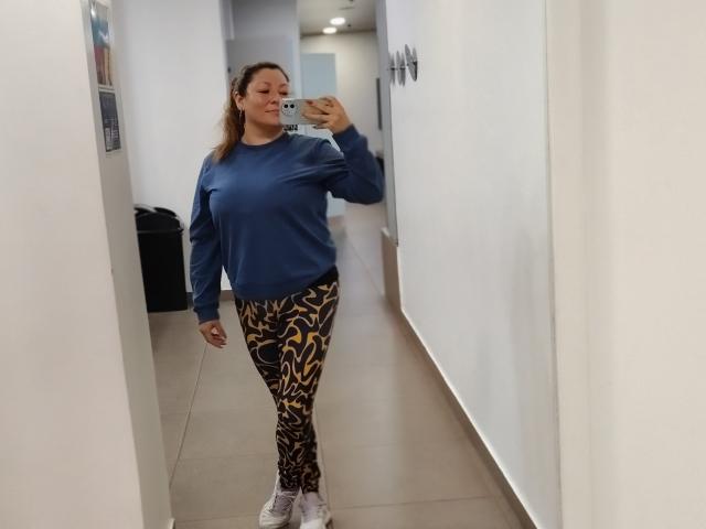 SakotoDika met bruin haar, een blauwe trui en gestreepte leggings in geel en zwart, staat in een gang en maakt een spiegelzelfportret. Ze draagt witte sneakers en houdt een telefoon vast..
