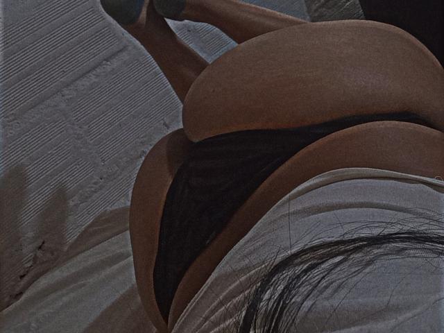El trasero de AmberMedellin es prominente, lleva ropa interior de encaje negro, está acostada en una cama con la espalda hacia la cámara, las piernas levantadas, una camiseta blanca parcialmente visible y su cabello oscuro se ve en el encuadre..
