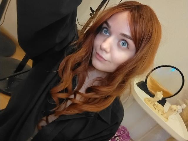 JanetShy with red hair, blue eyes, and pale skin, wearing a black shirt, selfie, mirror on right, wooden floor, black chair, and pink flowers in the background.