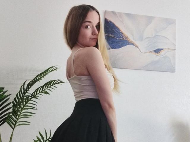 JanetShy with long brown hair, white tank top, and black skirt, looking back over her shoulder, showing her ass, standing next to a green plant and white wall.