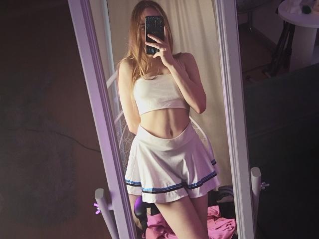 JanetShy with blonde hair, wearing a white crop top and skirt, holding a phone in front of her face, showing her toned midriff and ass. Mirror selfie.