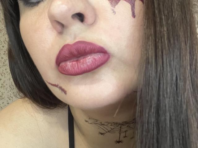 AlinaMilf's face with dark brown hair, red lipstick, and purple makeup stains on her cheeks and chin. She has a black spider tattoo below her neck.