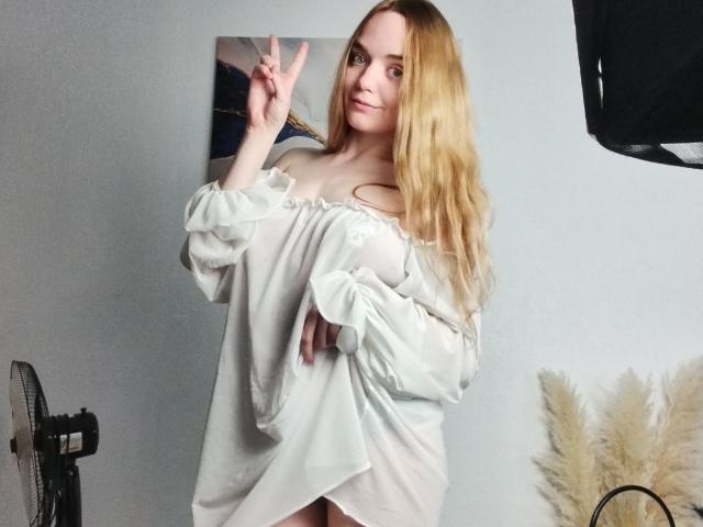 JanetShy with long blonde hair, off-shoulder white dress, showing a peace sign, smiling, standing, holding dress slightly up, revealing her bare ass.
