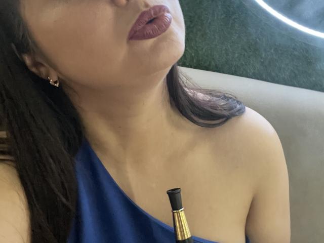 AlinaMilf mit dunklem Lippenstift, einem blauen Top mit einem Schulterträger, goldenen Ohrringen und einem Lippenstifthalt auf ihrer Brust, wobei ihre Brüste leicht sichtbar sind. Ihr Gesicht ist leicht nach oben geneigt..