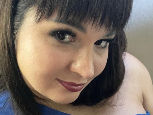 A AlinaMilf tem olhos castanho-escuros, sobrancelhas grossas e está usando batom vermelho. Ela está sorrindo, e seus seios mal são visíveis em uma blusa azul. Seu cabelo castanho-escuro tem franja..
